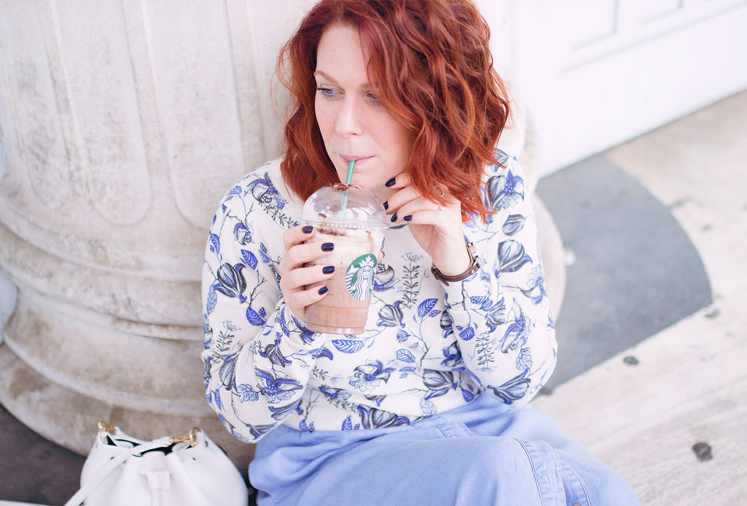 Assise sur le sol en damier contre une colonne avec une jupe midi en jean, un pull à fleurs, des derbies vernis bleu et un Starbucks pour la rentrée