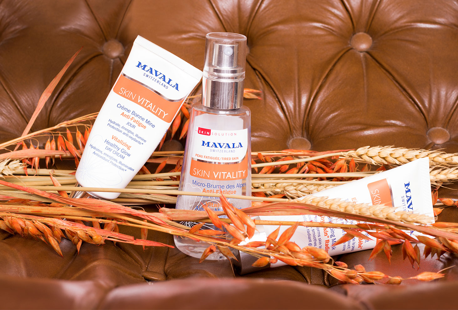 Trois produits de la gamme Skin Vitality par Mavala sur un fauteuil en vieux cuir marron au milieu de fleurs sèches oranges