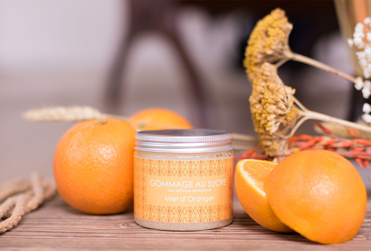 Le gommage au sucre Miel d'Oranger de L'art du Bain sur une planche en bois au milieu des oranges entières et de quelques fleurs sèches