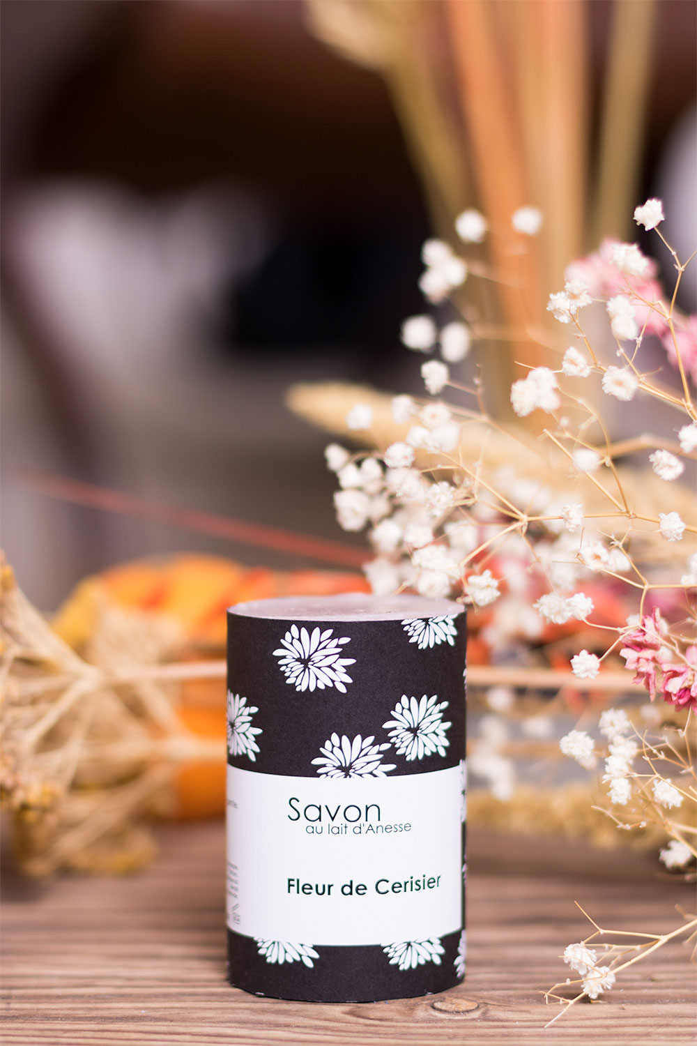 La packaging fleuri noir et blanc du savon Sento de L'art du Bain posé sur une planche en bois au milieu des fleurs sèches