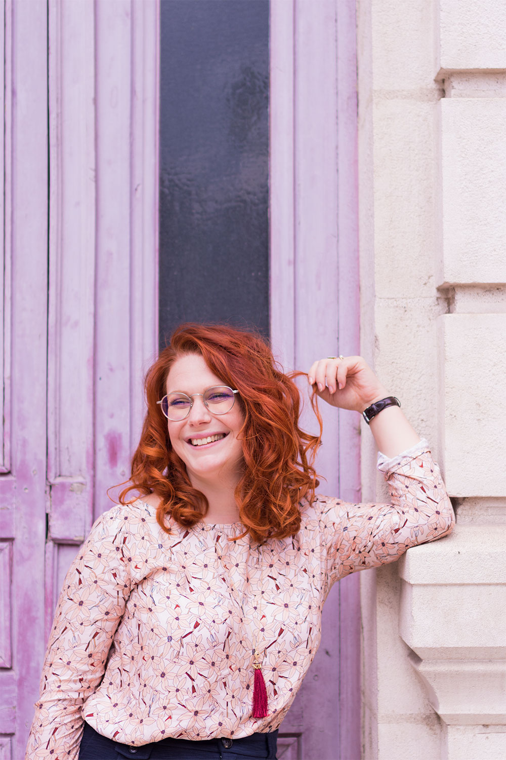 Zoom sur la blouse rose fleurie de la marque française La Morue, portée devant une porte rose fushia avec le sourire et la main dans les cheveux