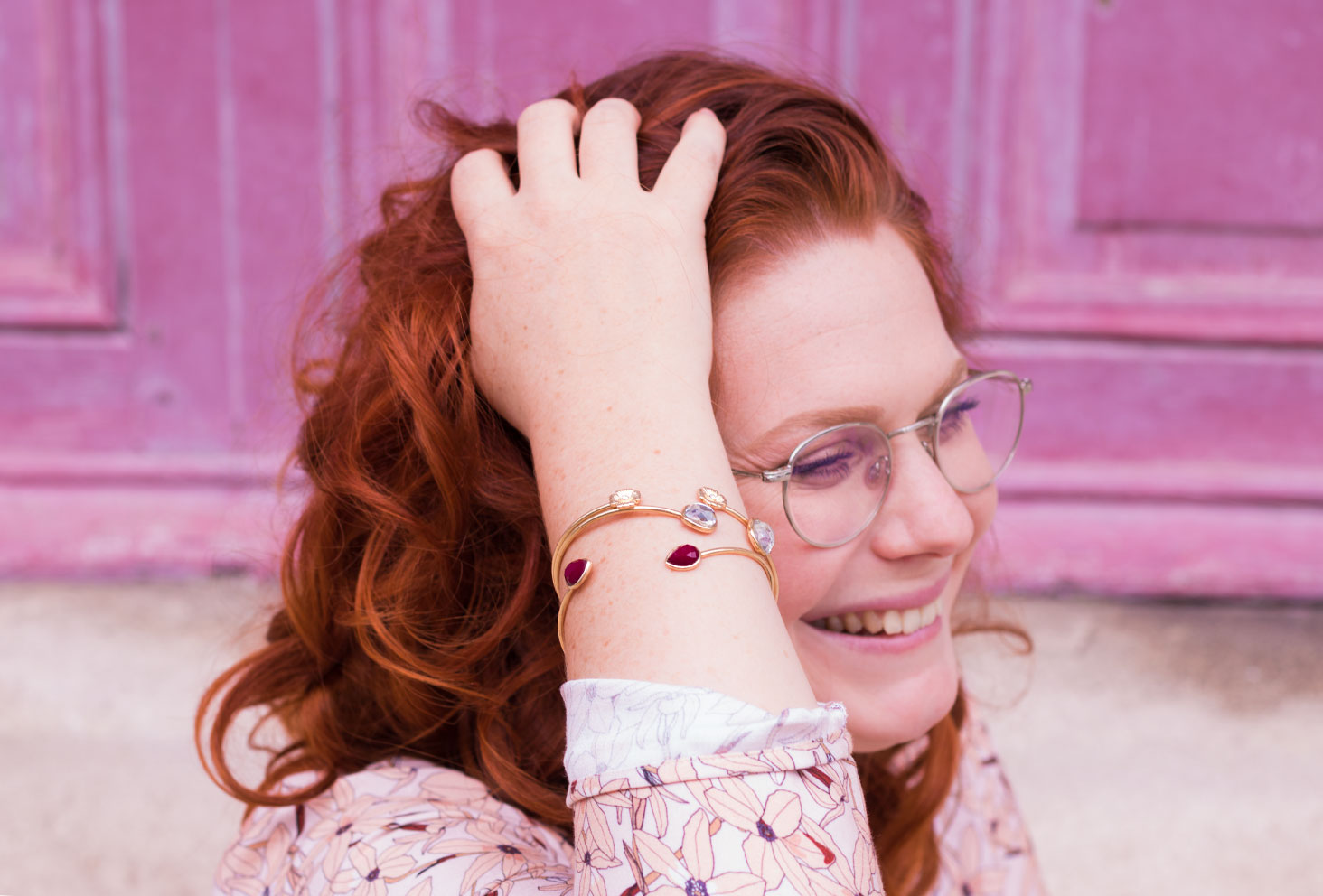 La main dans les cheveux, devant une porte rose, zoom sur les bracelets dorés MOA Paris