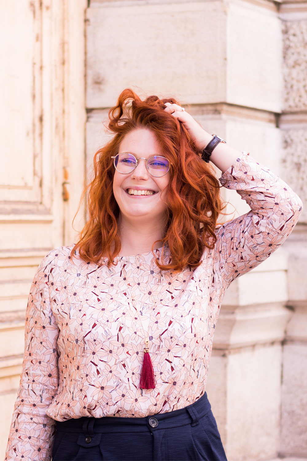 Portrait en blouse La Morue rose à fleurs, sautoir à pompon MOA, la main dans les cheveux bouclés et le sourire aux lèvres