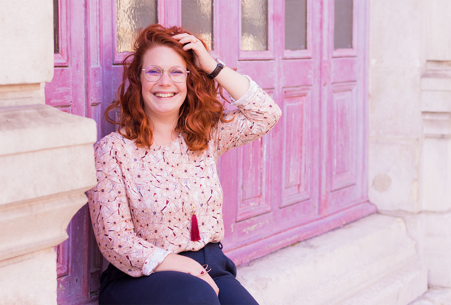 Assise le long d'une porte en bois fushia, la main dans les cheveux et le sourire aux lèvres, dans une blouse rose La Morue