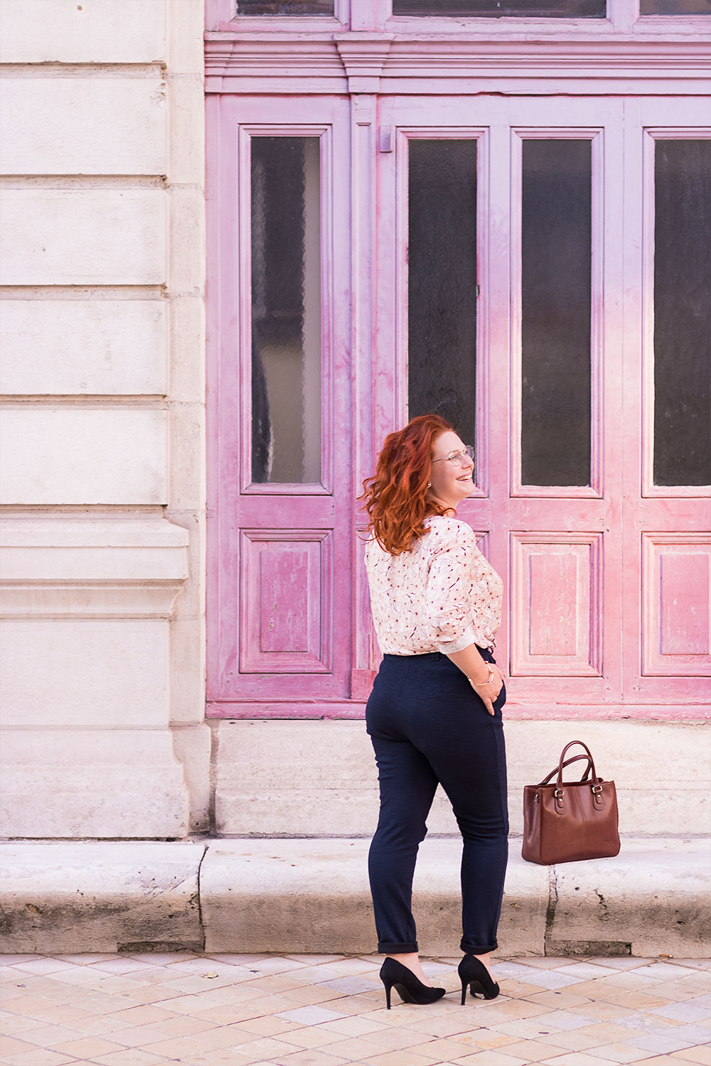 En entière de dos, devant les portes roses du grand théâtre de Tours, sur des escarpins hauts noirs et pantalon cigarette bleu marine, le sac posé aux pieds