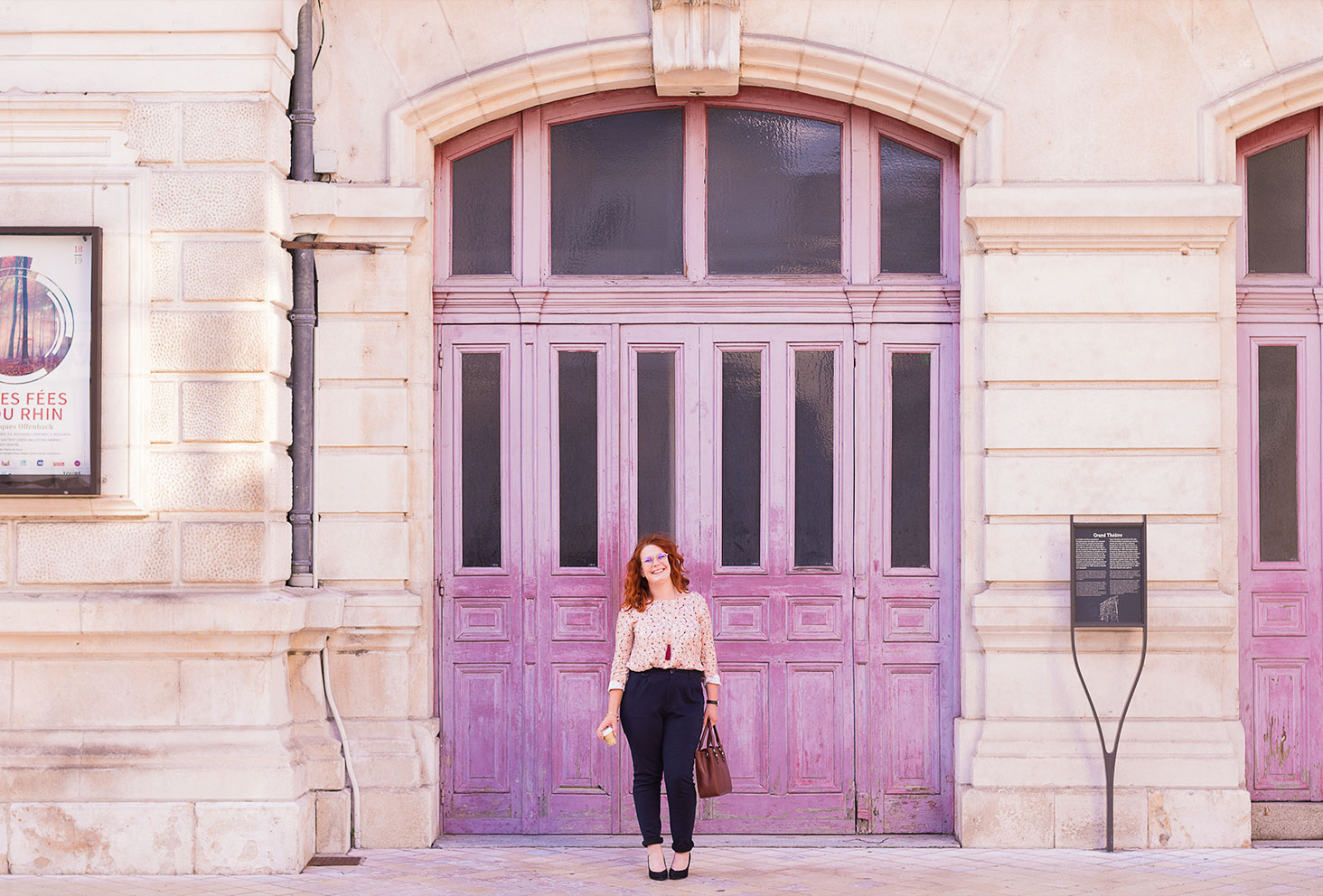 En look de working girl, pantalon cigarette et escarpins à talons hauts, devant les grandes portes en bois roses de l'opéra de Tours