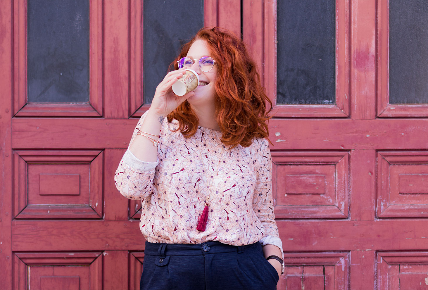 Un café à emporter à la bouche, en blouse rose à fleurs La Morue et les cheveux bouclés, devant une porte en bois rouge orangé