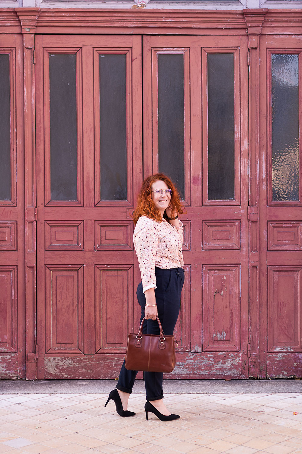 De profil devant une vieille porte en bois orange, en style working girl, blouse fluide La Morue et pantalon cigarette confortable, talons et sac en cuir dans la main