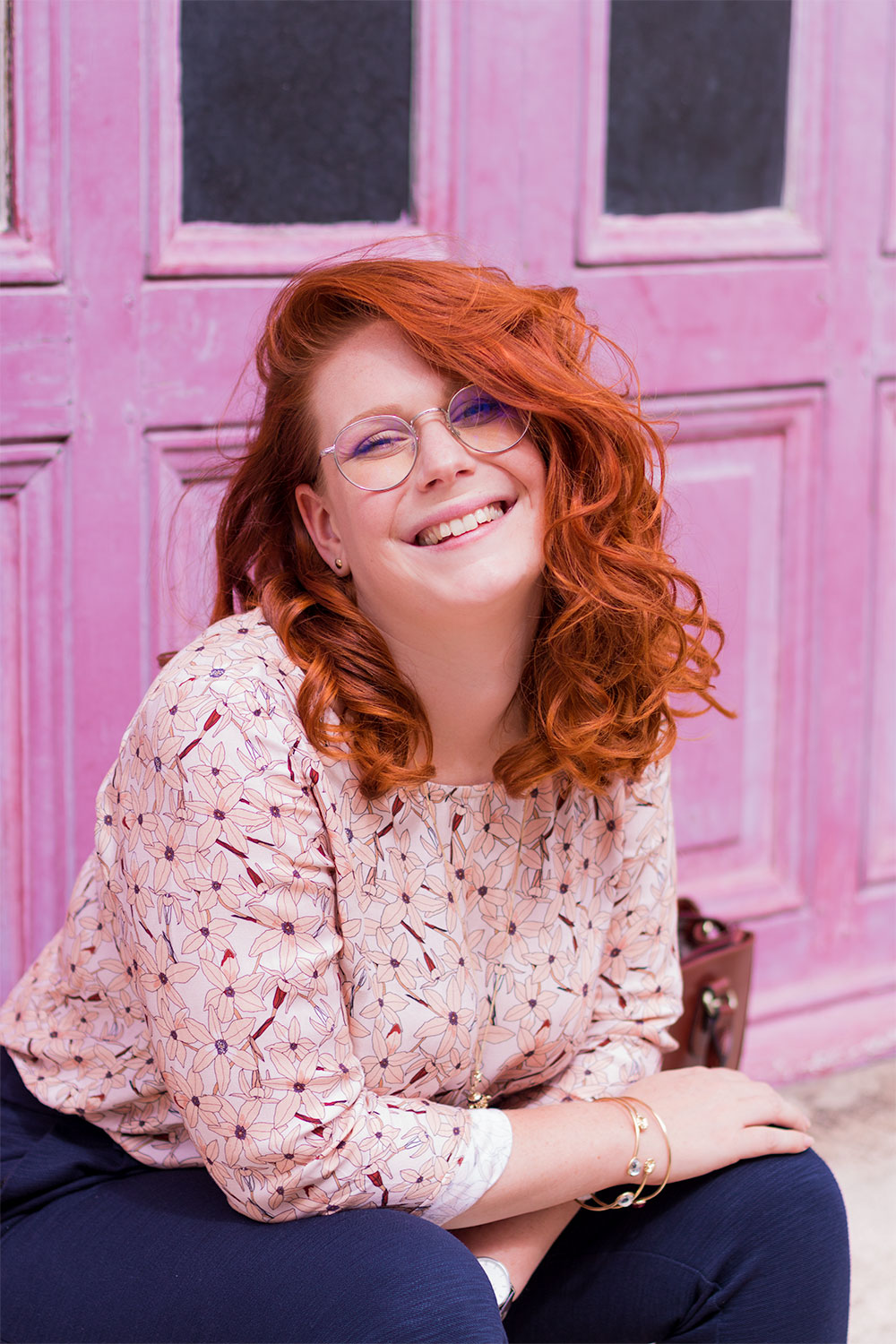 Portrait en blouse fluide rose La Morue, les cheveux énormément bouclés, devant une porte en bois rose fushia