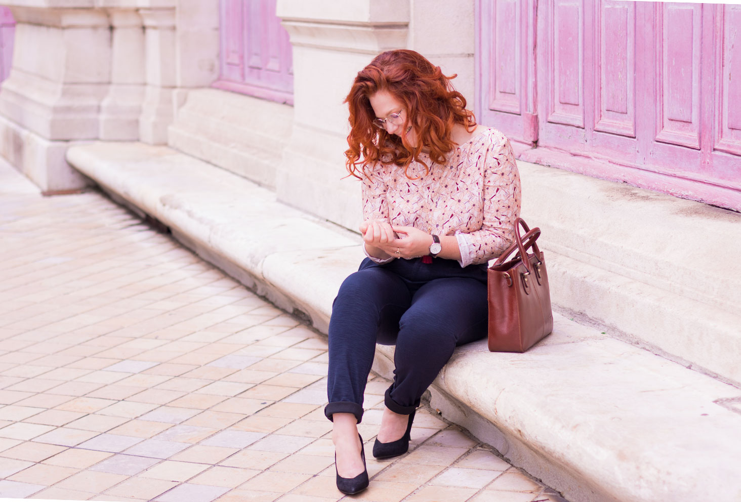 Assise de profil sur des marches en pierre devant deux grandes portes en bois fushia, en style working girl blouse et pantalon cigarette