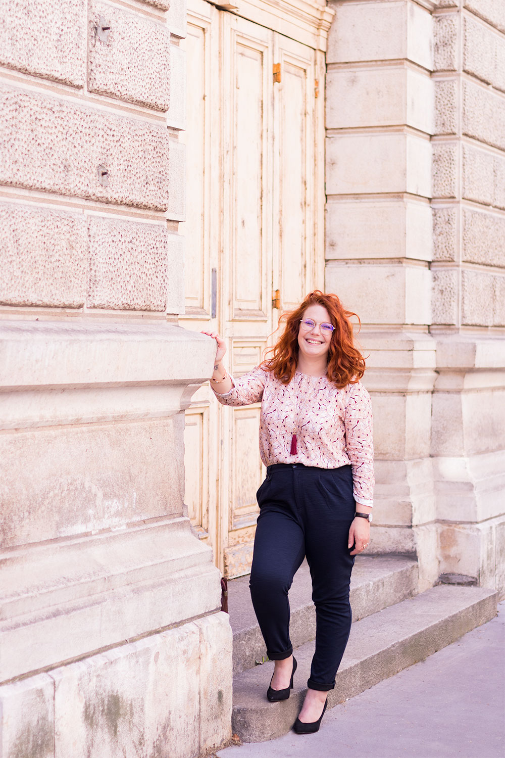 En working girl, blouse à fleurs roses et pantalon cigarette, le long d'un grand bâtiment en pierres blanches