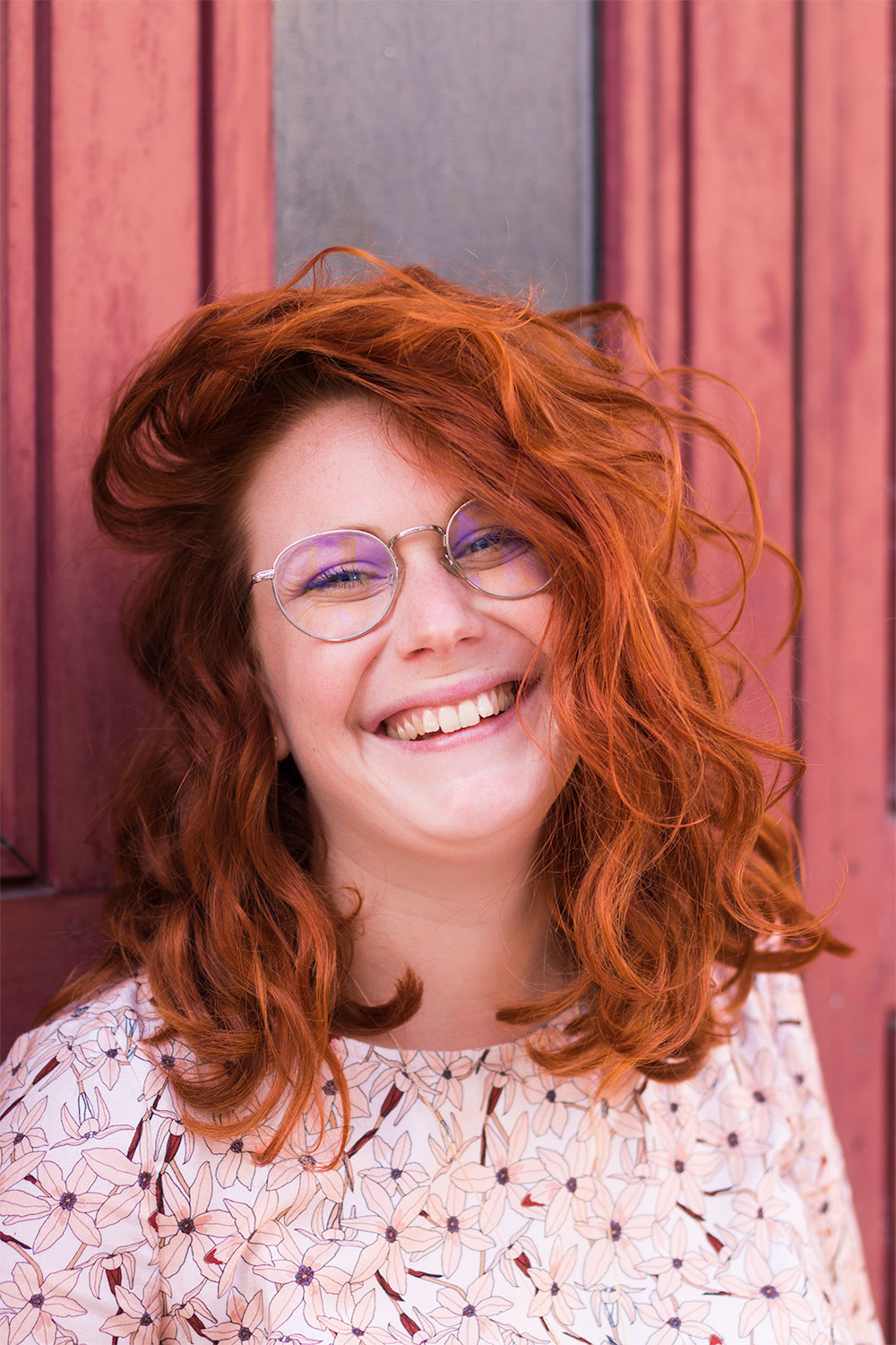 Photo portait serré, cheveux roux énormément bouclés, avec un grand sourire aux lèvres et des yeux bleus cachés derrière de grosses lunettes de vue