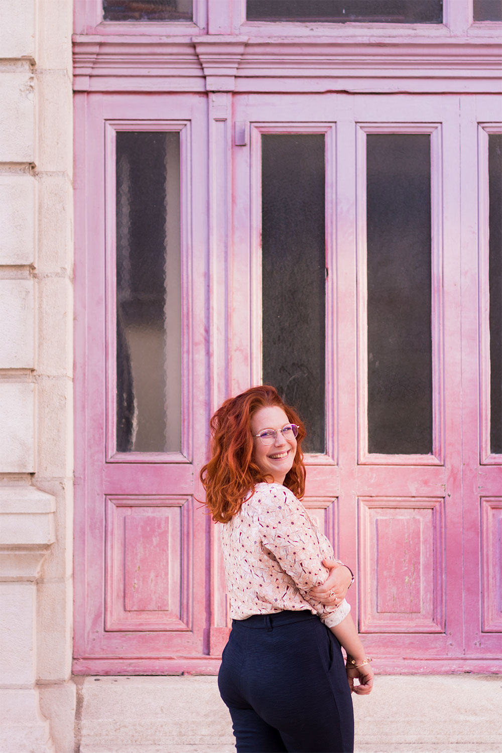 De dos, devant une porte en bois rose, en blouse La Morue et pantalon confortable bleu marine