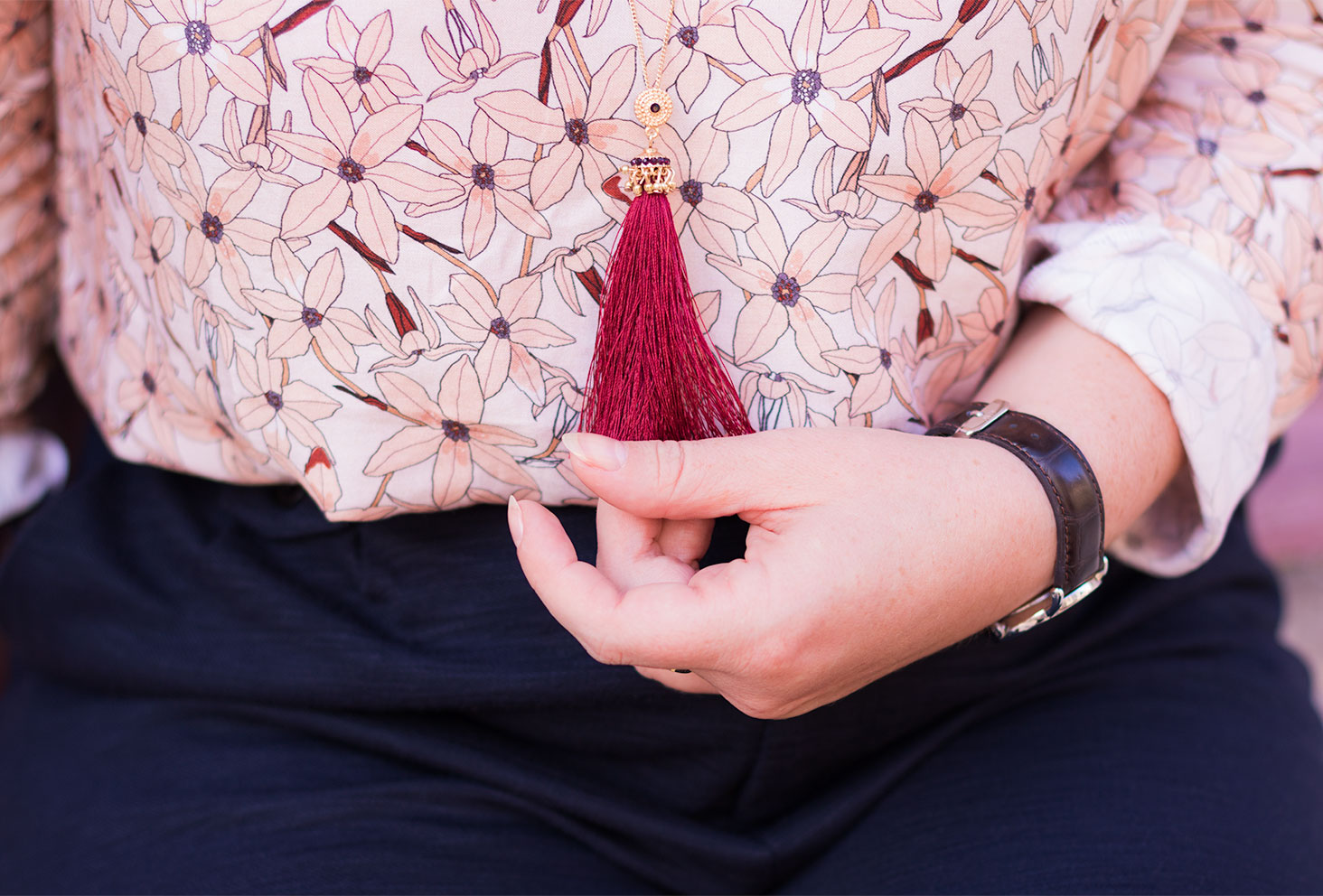 Zoom sur le pompon du sautoir MOA bordeaux portés et tenu dans une main devant la blouse fleurie La Morue