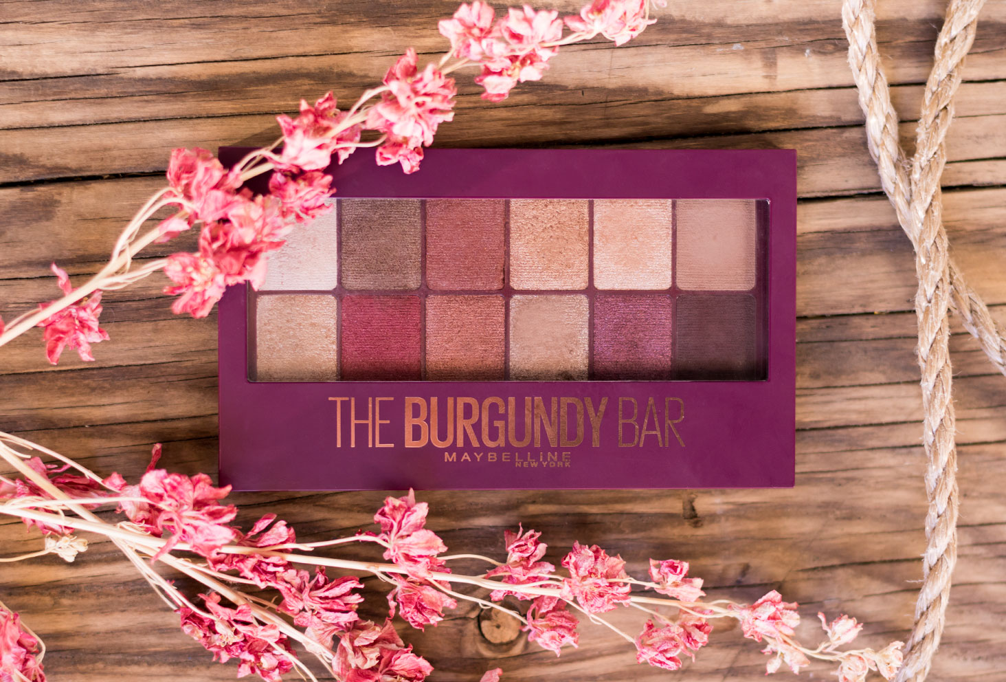 La palette The Burgundy Bar de haut fermée, sur une table en bois, au milieu de cordages et de fleurs sèches roses