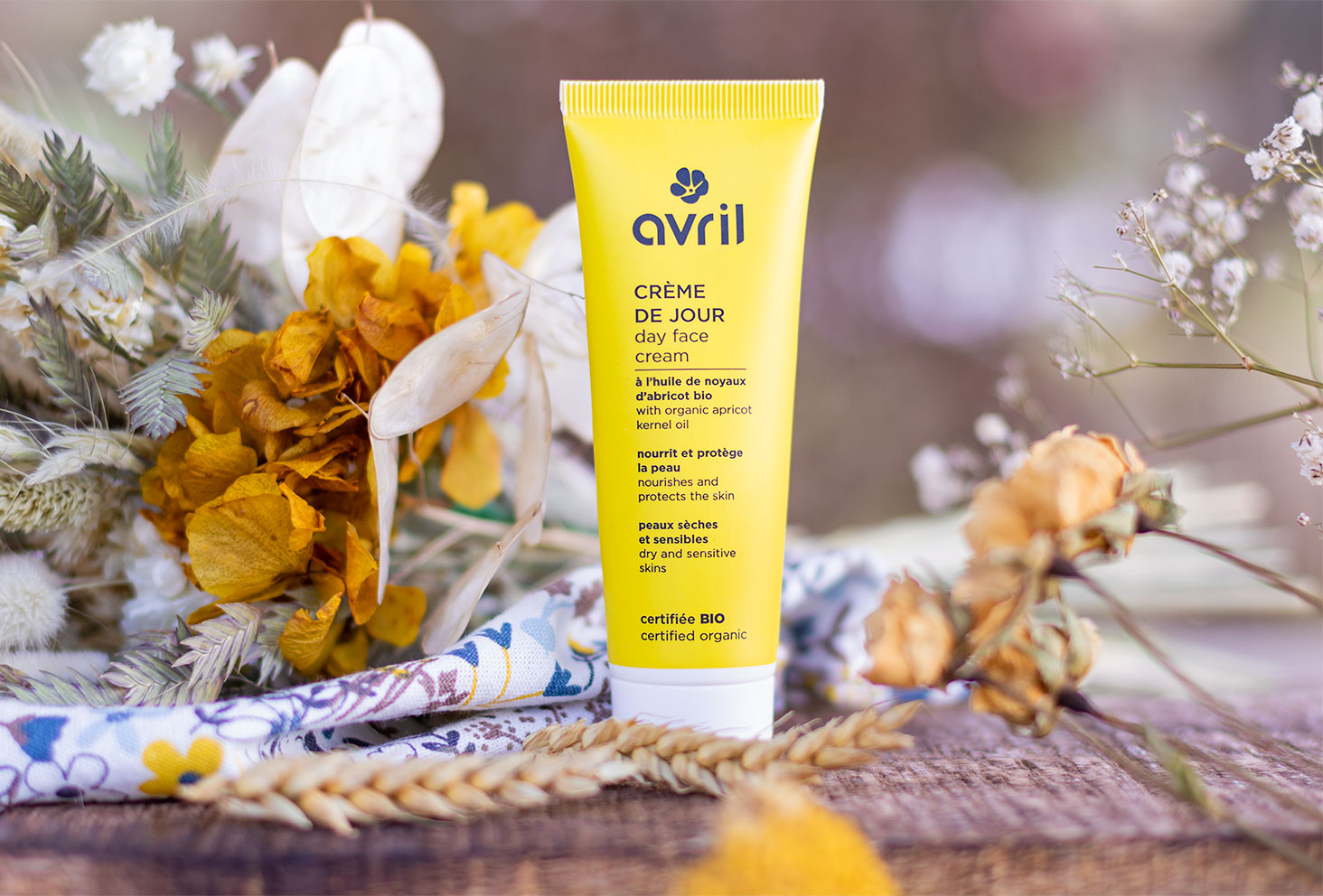 La crème de jour au packaging jaune de la marque Avril, debout sur une planche en bois à côté d'un bouquet de fleurs séchées jaunes et blanches