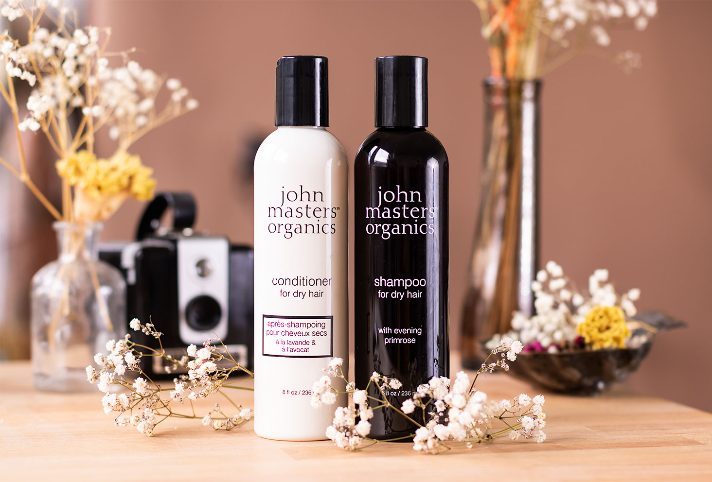 Le shampooing et l'après-shampooing pour cheveux secs de la marques John Masters Organics, posés sur un bureau au milieu de fleurs sèches et de décorations vintages