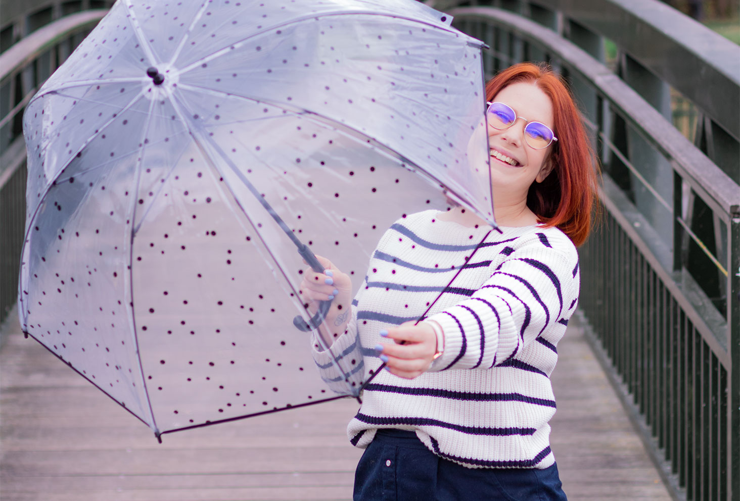 Cachée derrière un parapluie à pois transparent tenu dans les mains, avec le sourire sur un pont en bois