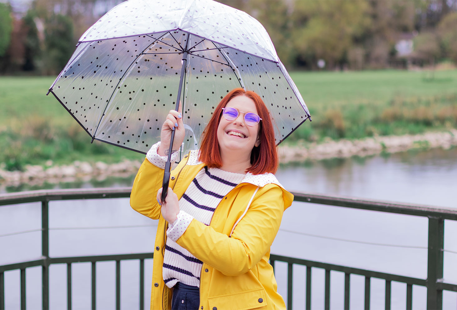 Sur un pont au bord de l'eau, en ciré jaune avec un parapluie transparent à motifs pois au-dessus de la tête