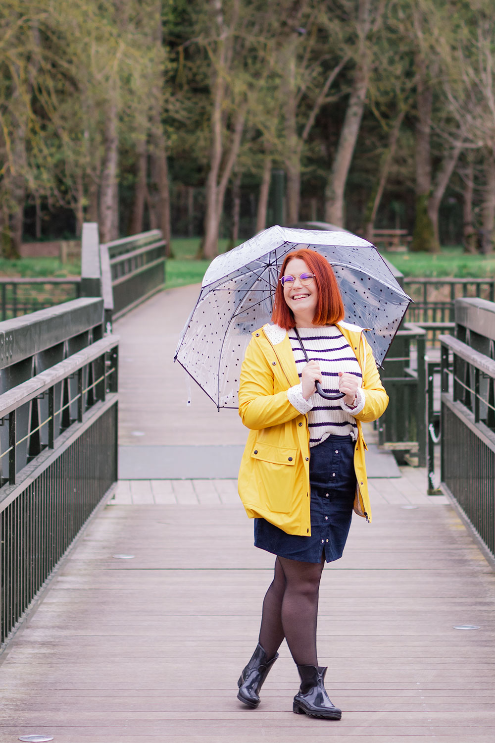 Au milieu d'un pont au-dessus de l'eau, en ciré jaune avec un parapluie dans la main, en pull marin sous la pluie