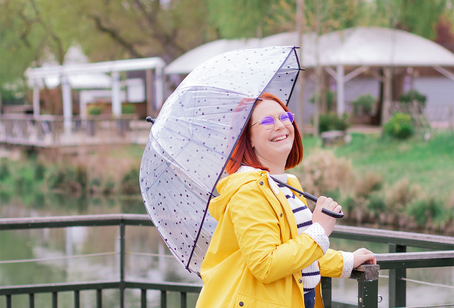 De profil, appuyée contre les rambardes d'un pont, en ciré jaune tenant un parapluie transparent posé sur l'épaule