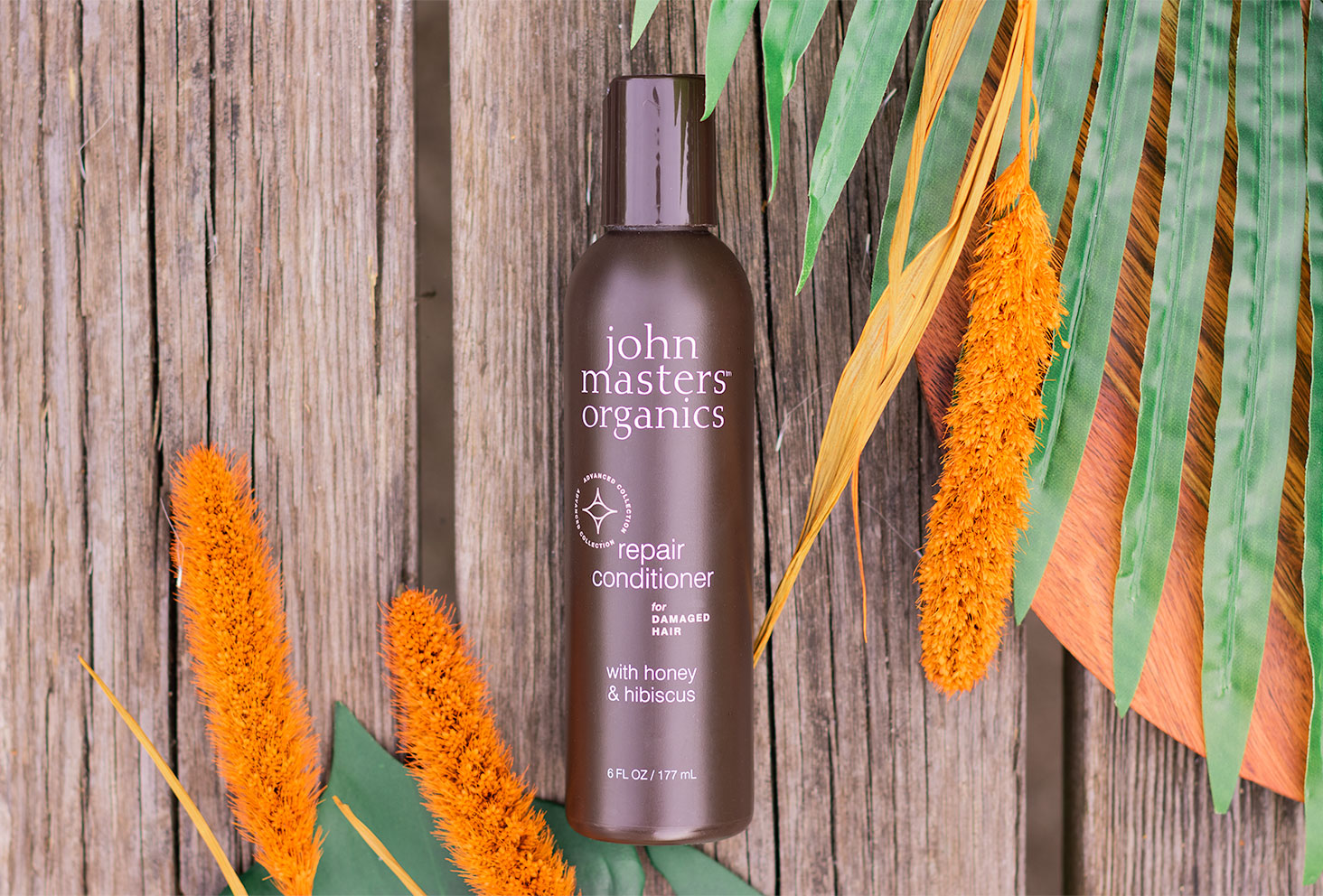 Zoom sur l'après-shampoing pour cheveux abîmés de John Masters Organics, allongé sur une table en bois au milieu de fleurs séchées