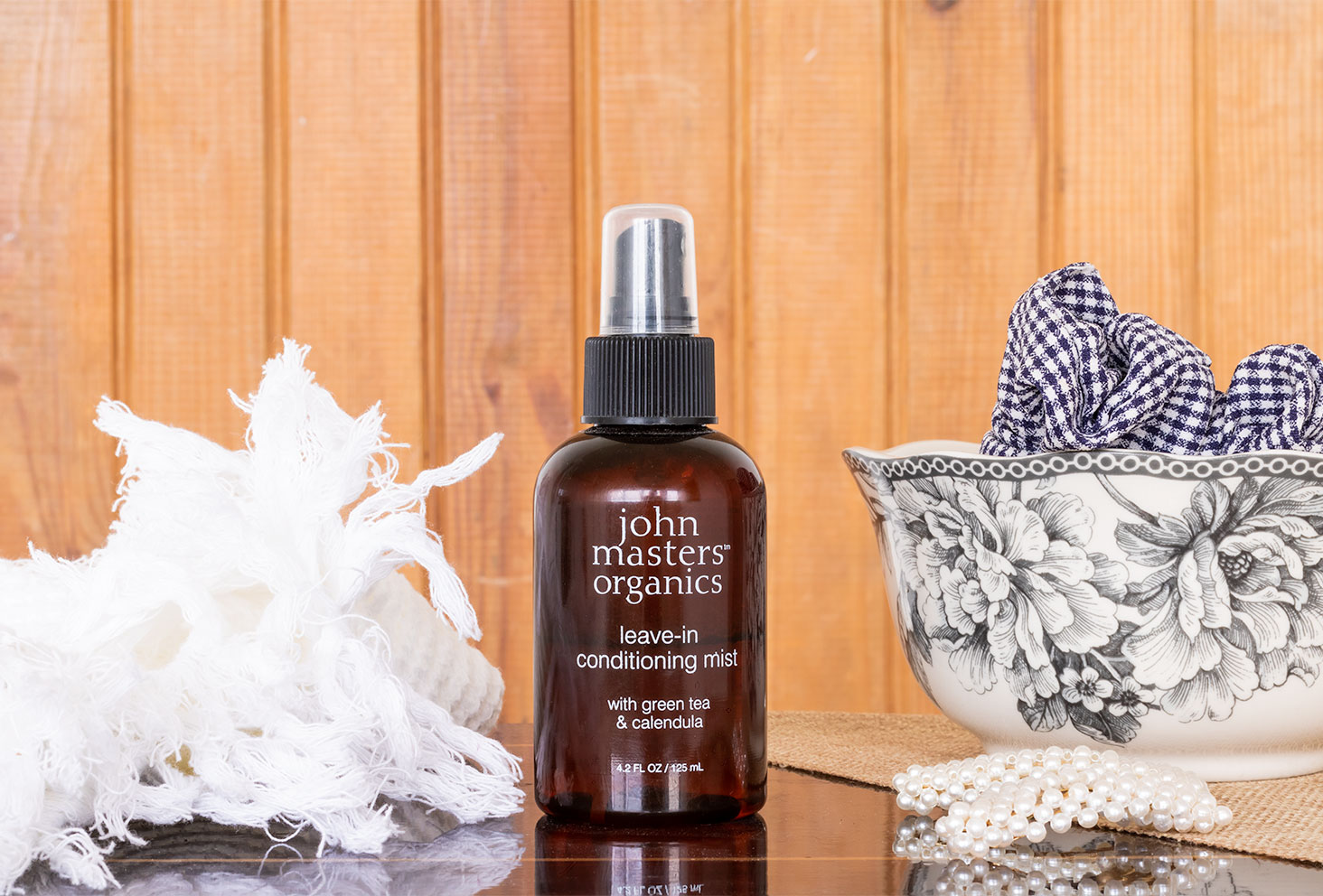 Zoom sur le spray revitalisant de John Masters Organics posé sur une table vernis au milieu d'accessoires cheveux dans une salle de bain en lambris