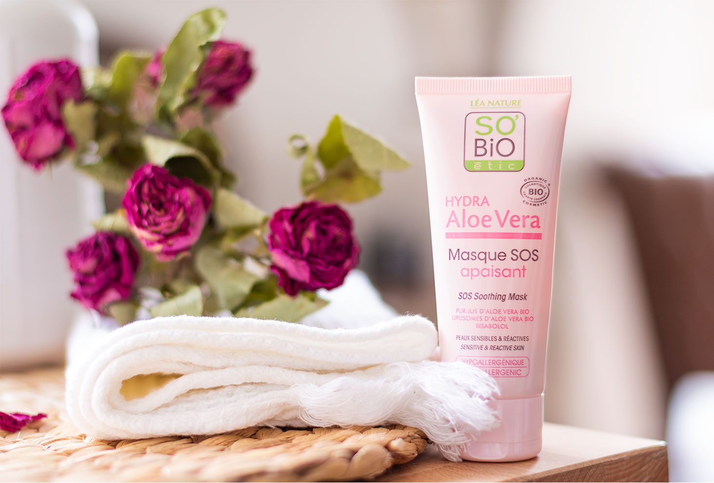 Le masque rose SOS apaisant à l'aloe vera de SO'BiO étic posé sur le coin d'un meuble de salle de bain, à côté de roses séchées et d'une serviette blanche sur un dessous en osier