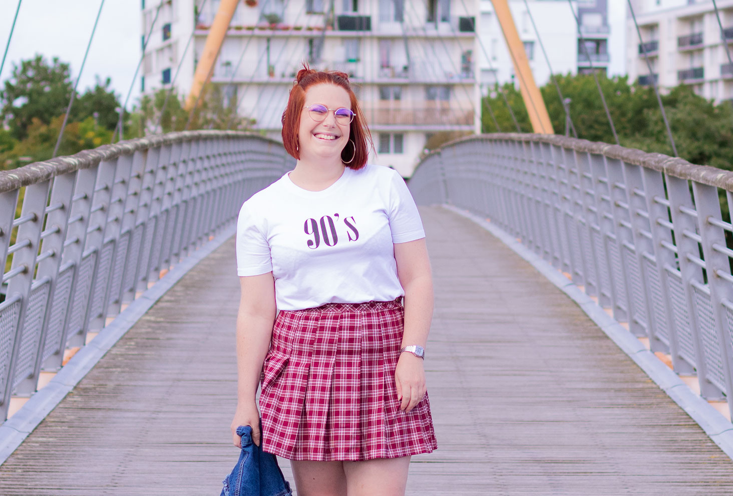 Au milieu d'un pont avec le sourire, dans un look vintage t-shirt 90's et jupe à carreaux bordeaux