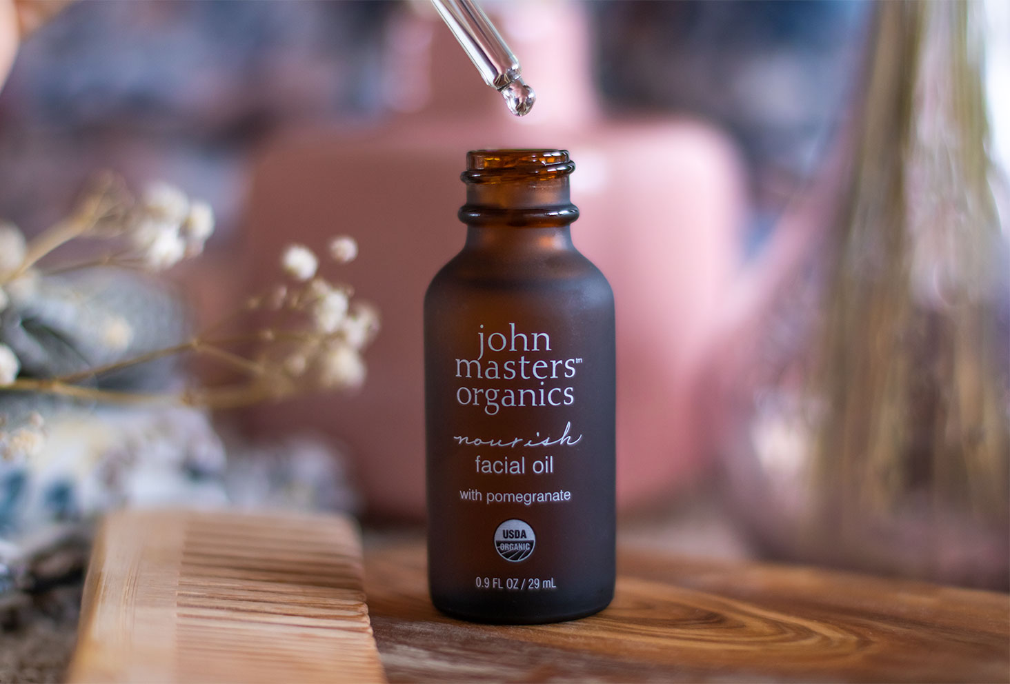 Utilisation du flacon en verre de l'huile nourrissante visage de John Masters Organics, posé sur une planche en bois avec sa pipette en verre tenue dans une main