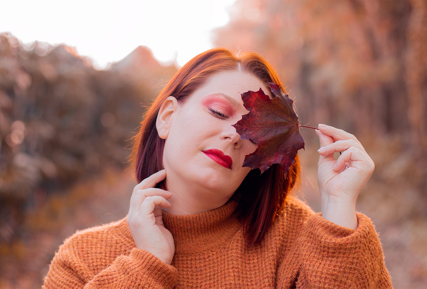 Zoom sur un maquillage aux couleurs de l'automne au milieu de la forêt, une feuille bordeaux tenue devant l'oeil gauche