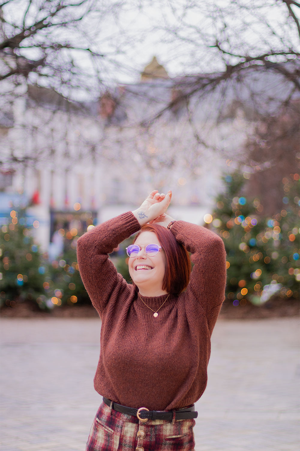 Les mains au-dessus de la tête avec le sourire, en pull en laine marron, devant les sapins décorés et allumés pour Noël