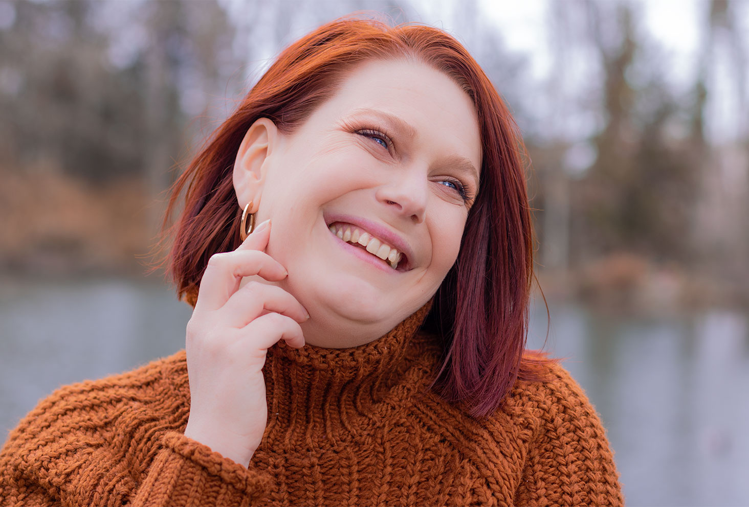 Portrait avec le sourire en pull marron à col cheminé, une main sur le visage, devant un lac