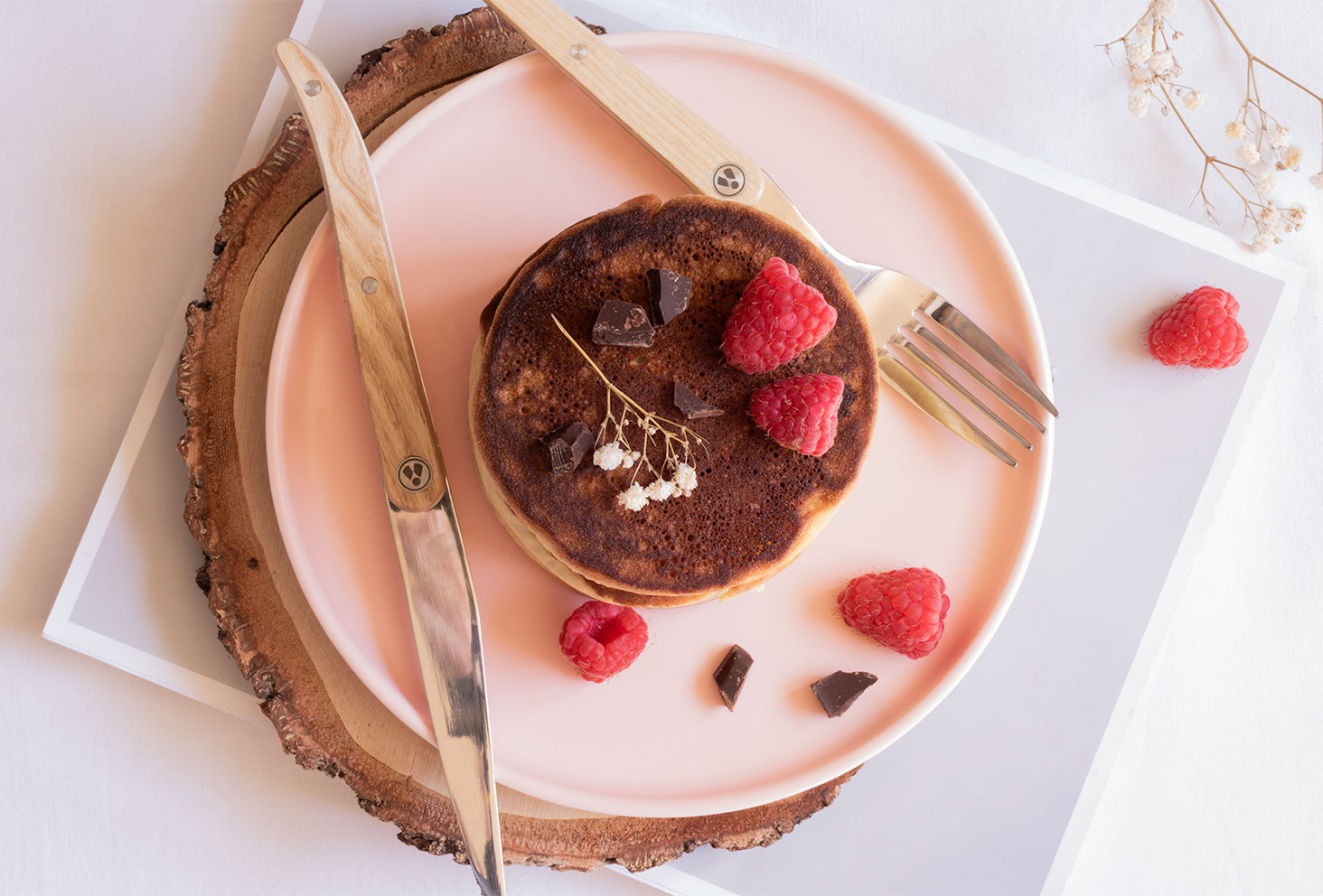 Une recette facile de fluffy pancakes, posés dans une assiette rose avec un couteau et une fourchette en bois, au milieu de framboises et d'éclats de chocolat