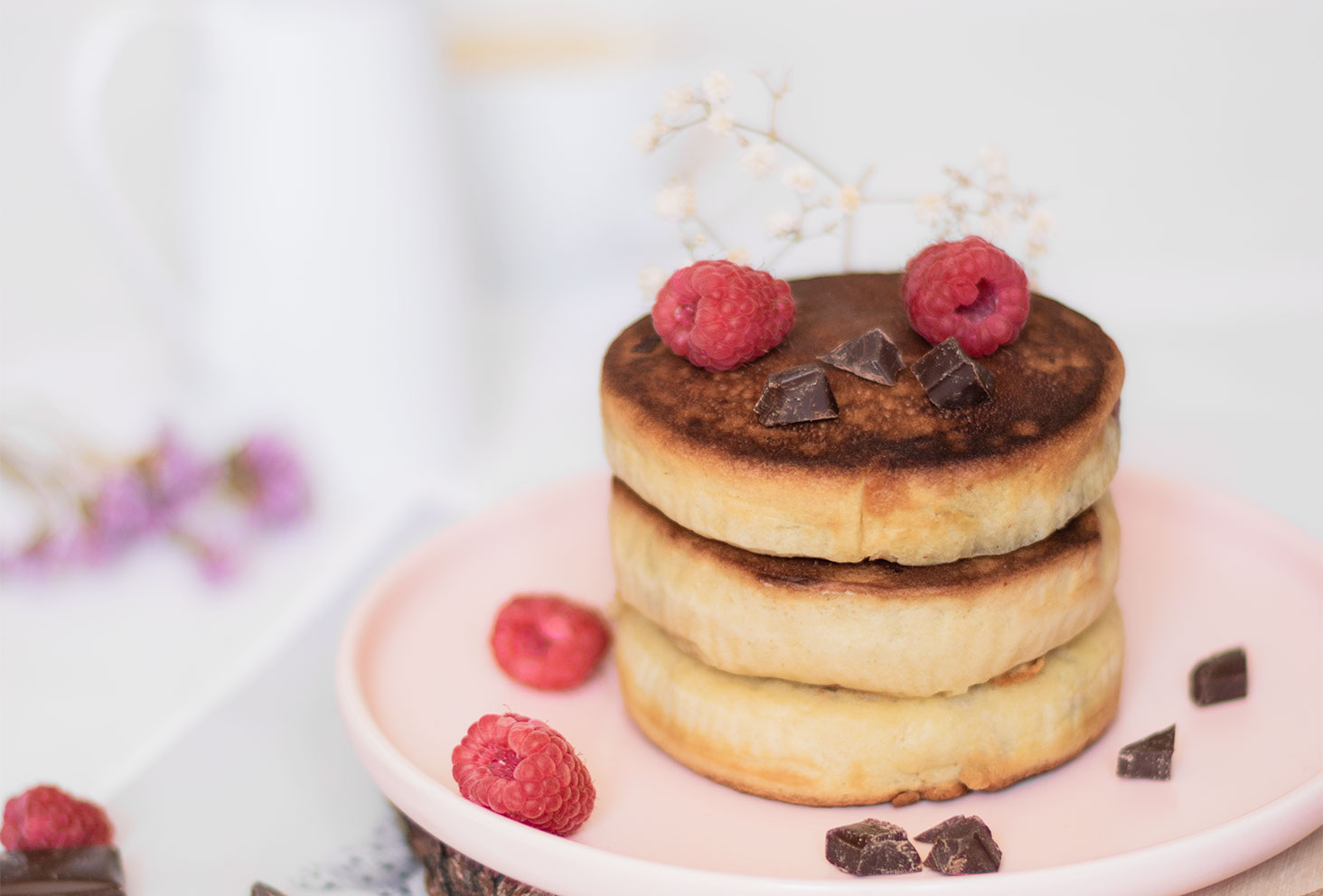 Zoom sur 3 pancakes fluffy dans une assiette rose au milieu de chocolat et de framboises