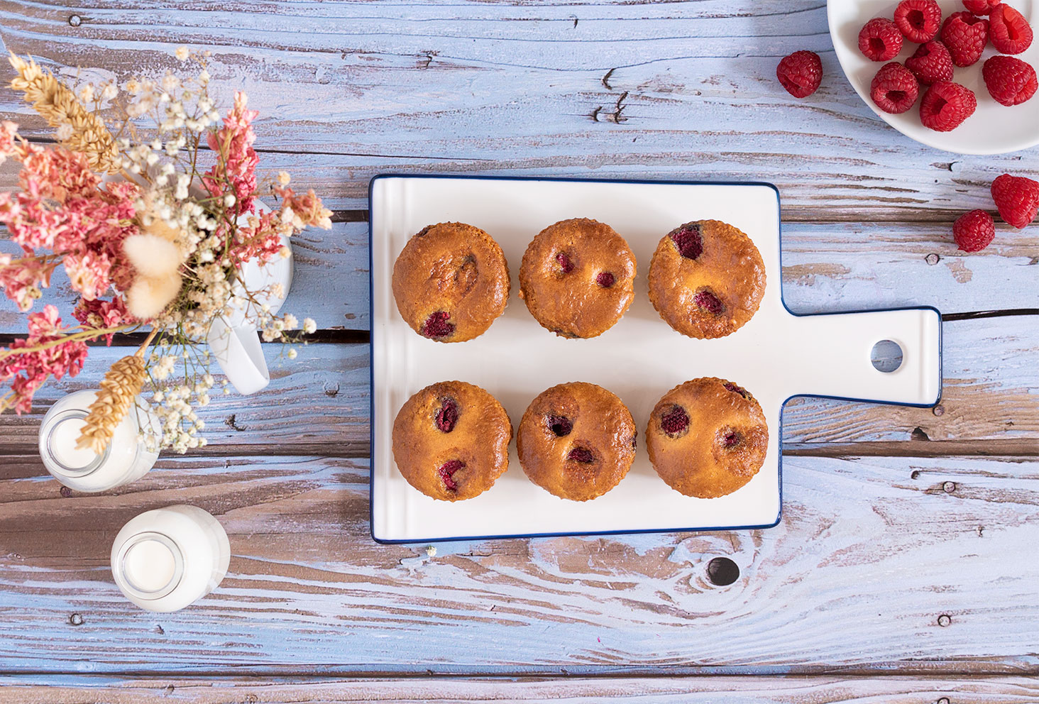 Des muffins aux framboises alignés sur une planche à découper en verre blanche, sur une table en bois bleutée