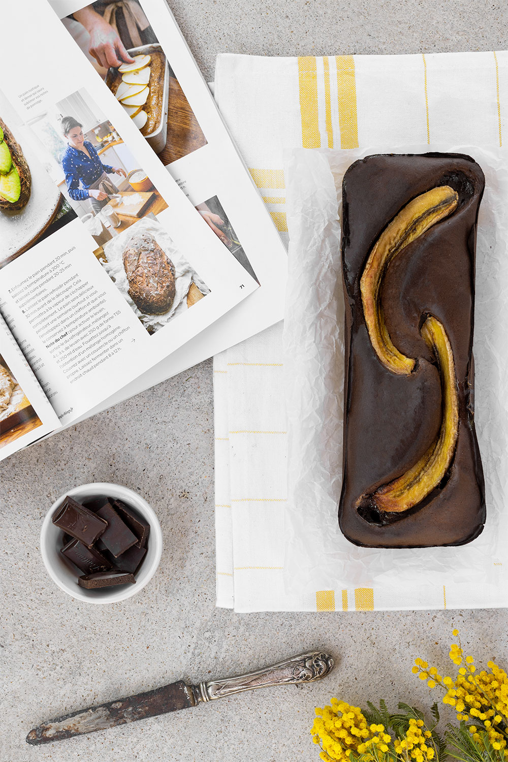 Un banana bread au chocolat protéiné posé sur un torchon à carreaux jaune et blanc, sur un fond en béton, à côté de fleurs jaunes et d'un magazine culinaire ouvert