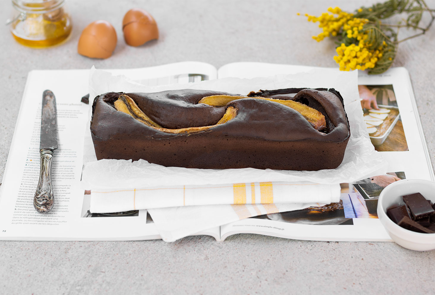 Un banana bread chocolat posé sur un magazine ouvert au milieu des ingrédients