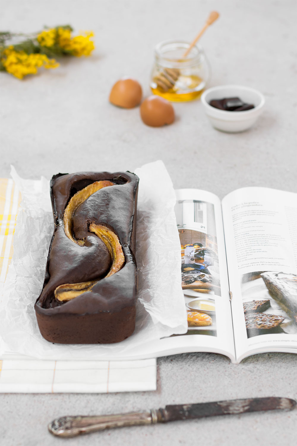 Recette facile et stylisme culinaire d'un banana bread au chocolat posé sur un magazine et un torchon à carreaux, au milieu d'ingrédients qui le compose