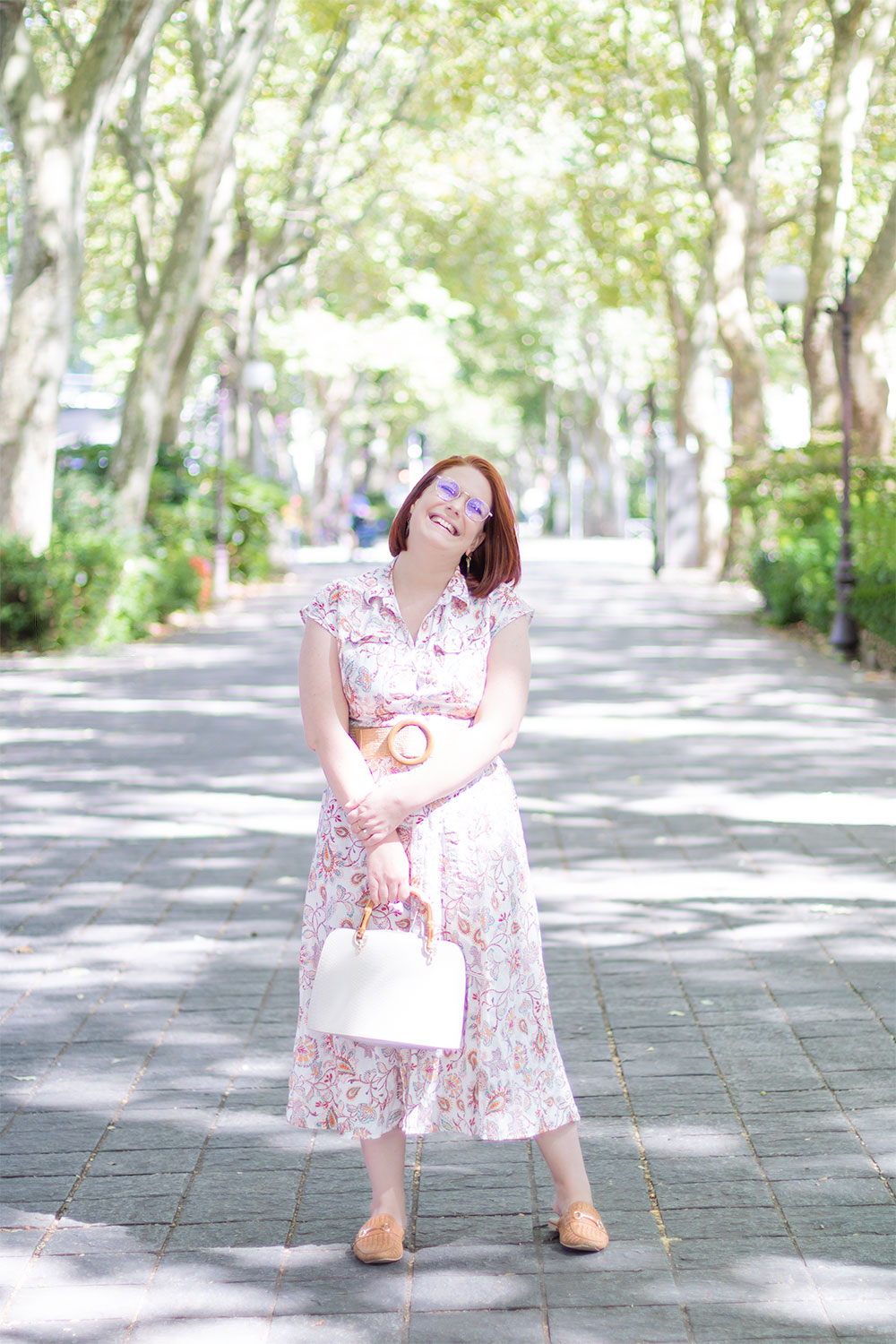 Au milieu du boulevard de Tours ensoleillée, en robe chemise longue fleurie et un sac beige dans les mains, avec le sourire