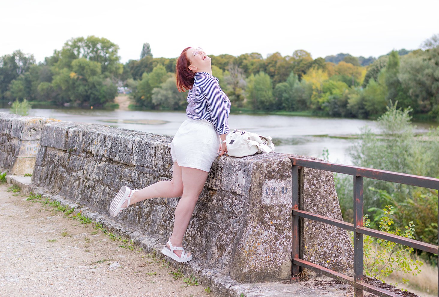 Appuyée sur le mur le long des bords de Loire, avec le sourire et un pied levé, en short et chemise rayée