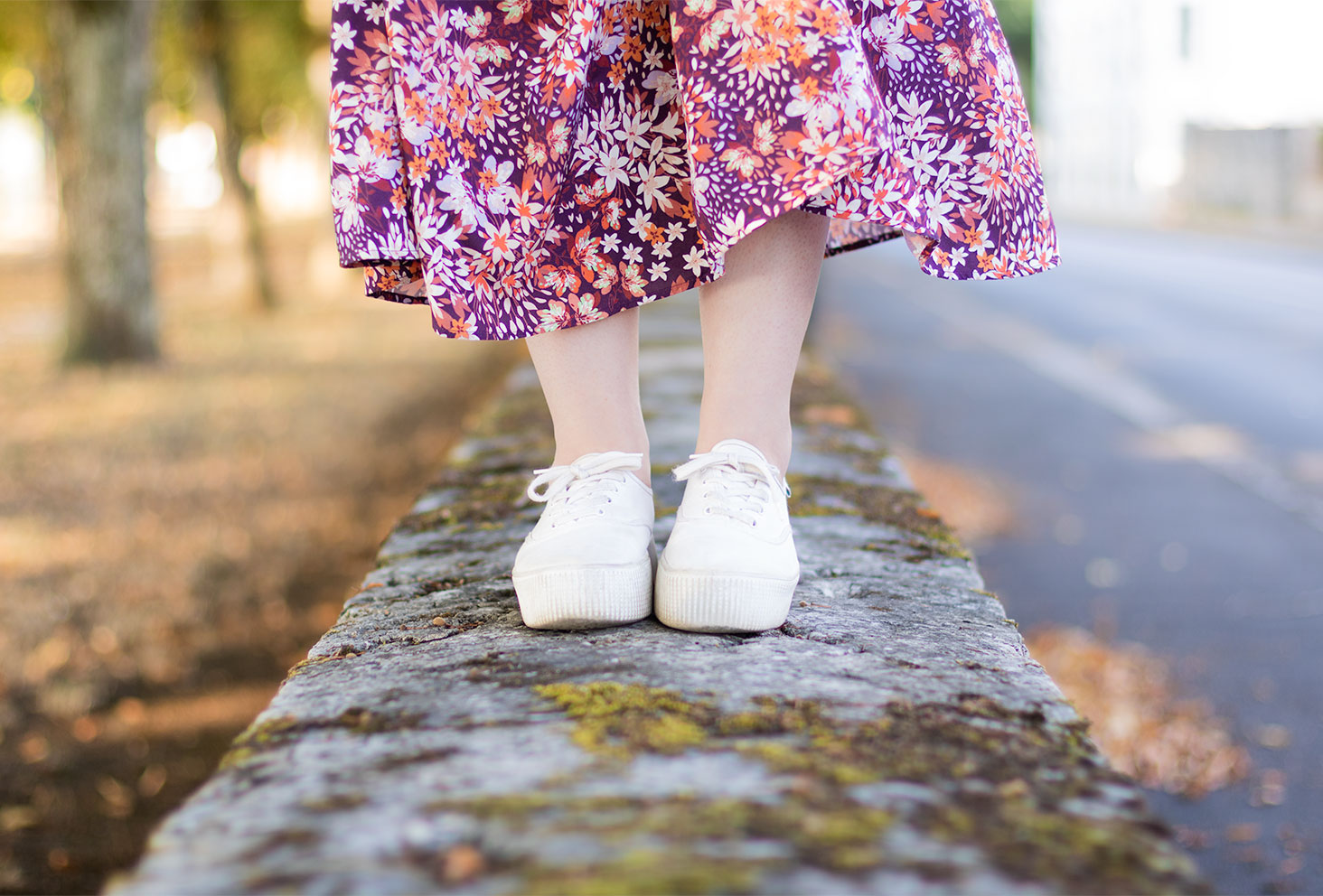 Zoom sur les baskets blanches Victoria portées avec une jupe longue rose, debout sur un mur en pierre