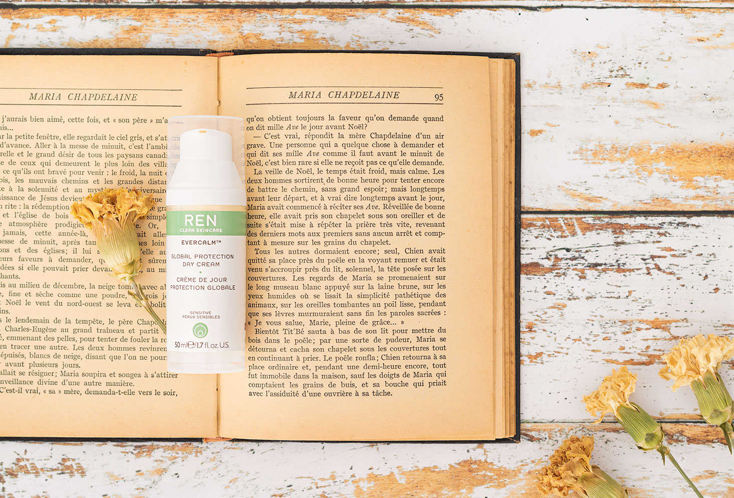 Zoom sur la crème de jour protection globale de Ren Clean Skincare posée sur un livre ancien ouvert avec une fleur séchée jaune