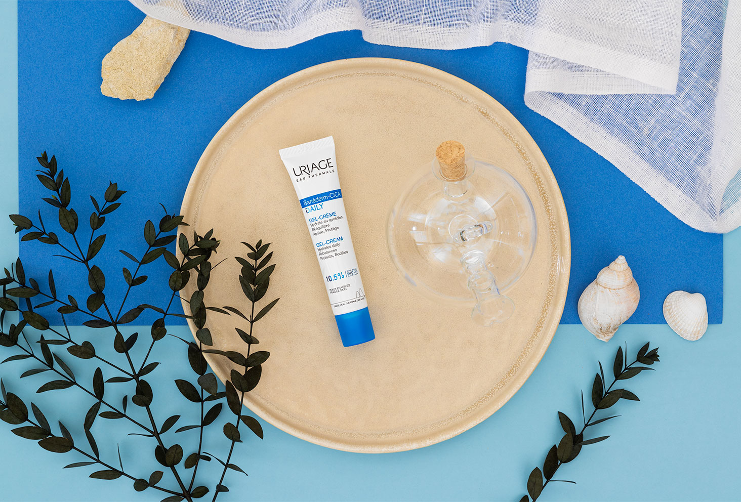 Le gel-crème hydratant et apaisant de la gamme Bariéderm-CICA d'Uriage dans une assiette ronde couleur sable, sur un fond bleu aux couleurs de la mer avec des accessoires d'été