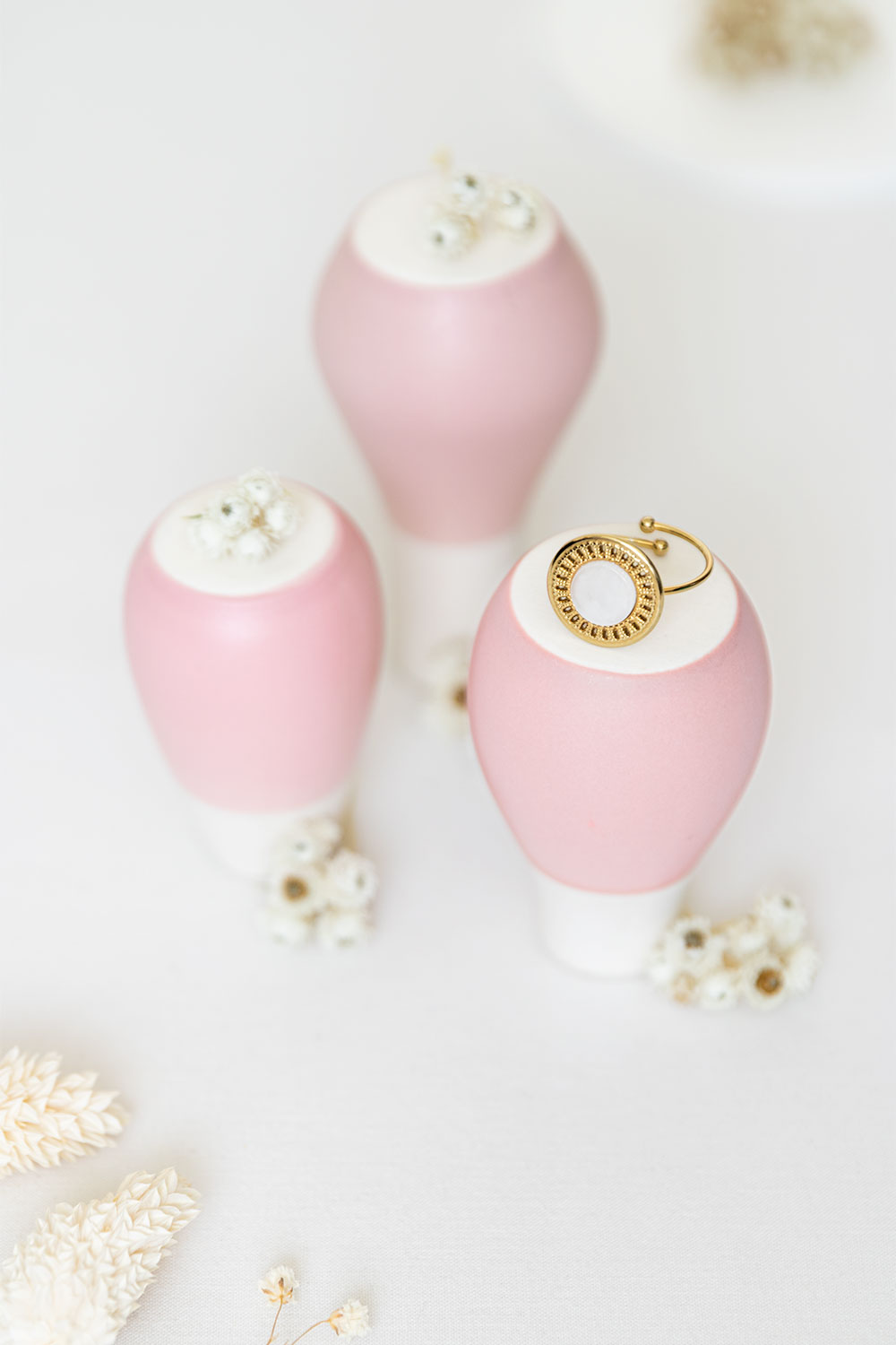 Bague dorée Amarok de la marque Vaniya Bijoux posée sur des vases rose poudré retrounés au milieu de fleurs séchées blanches