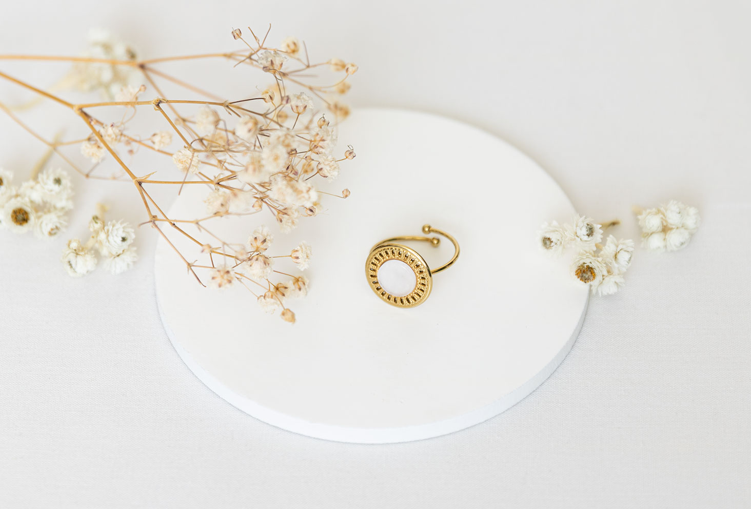 La bague dorée Vaniya Bijoux sur fond blanc, posée sur un support rond blanc au milieu de fleurs séchées