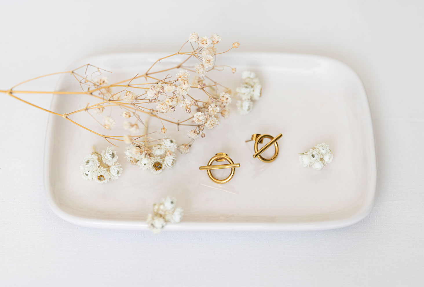 Les boucles d'oreille dorées Vaniya Bijoux sur fond blanc, posées dans une coupelle rectangulaire blanche au milieu de fleurs séchées