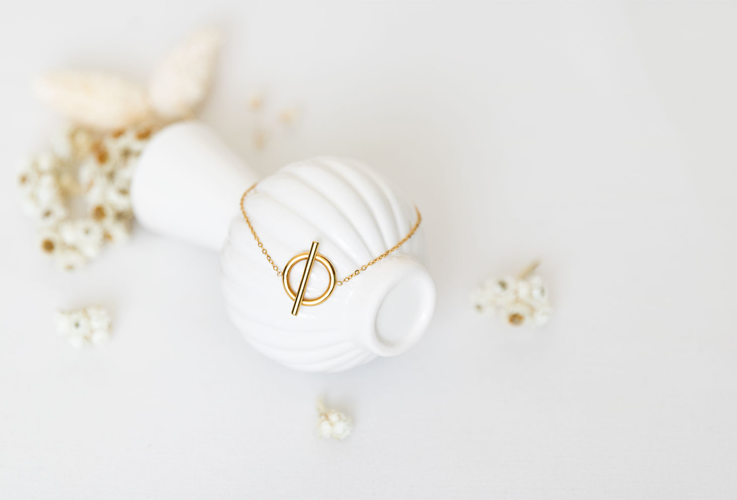 Le bracelet doré Vaniya Bijoux sur fond blanc, posé sur un vase renversé blanc au milieu de fleurs séchées