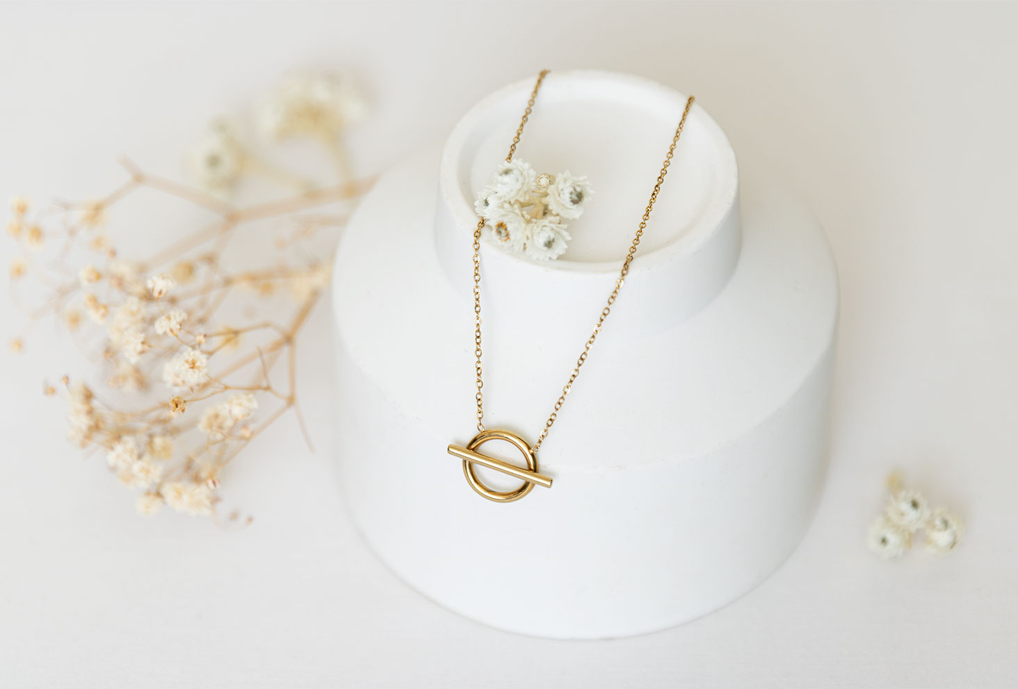Le collier doré Vaniya Bijoux sur fond blanc, posé sur un gros vase blanc au milieu de fleurs séchées