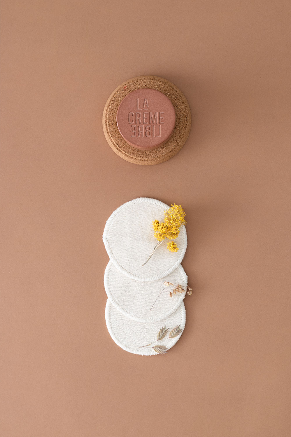 La crème riche de La Crème Libre dans un pot terracotta sur fond marron à coté de cotons lavables beiges